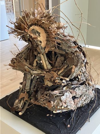 A sculpture made of natural materials such as branches, leaves, and bark. The figure appears to be sitting, with a crown of spiked wooden elements surrounding its head, blending organic textures into a humanoid form.