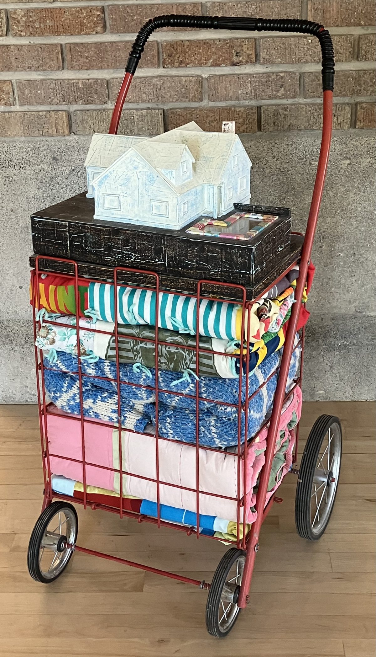 A red shopping cart filled with folded colorful quilts, topped with a model of a house. The house is painted light blue, and the structure rests on top of a black base that appears to be a weathered box.