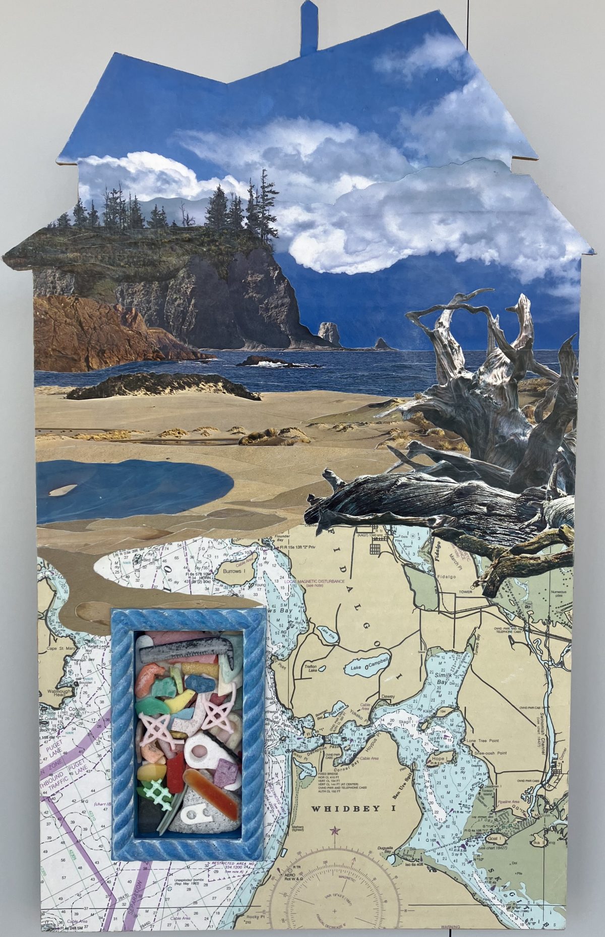 A house-shaped collage featuring a rocky coastline, beach, and cloudy sky. A section of a map is integrated into the lower part of the piece, with a small box in the center filled with colorful found objects.