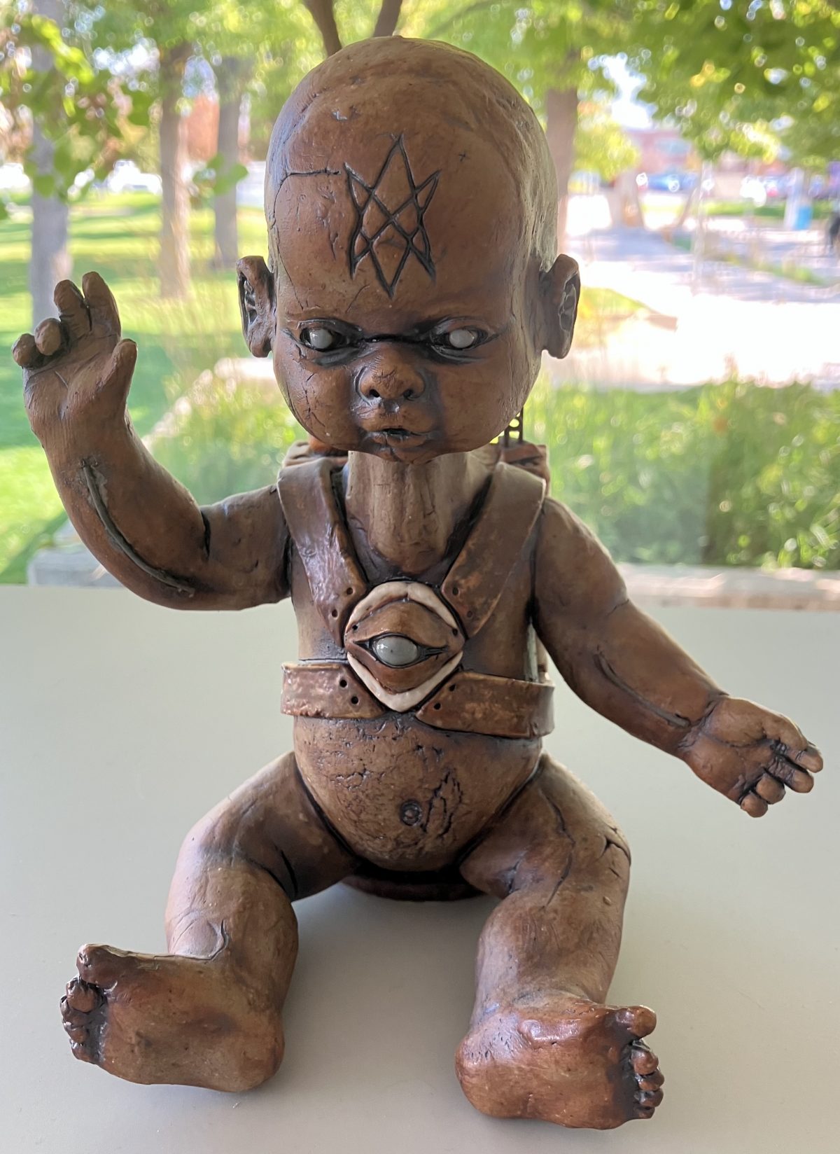 A small sculpture of a baby, with an unsettling expression and geometric markings on its head. The figure is seated, one arm raised, wearing a harness with an eye-like emblem at the center of its chest.