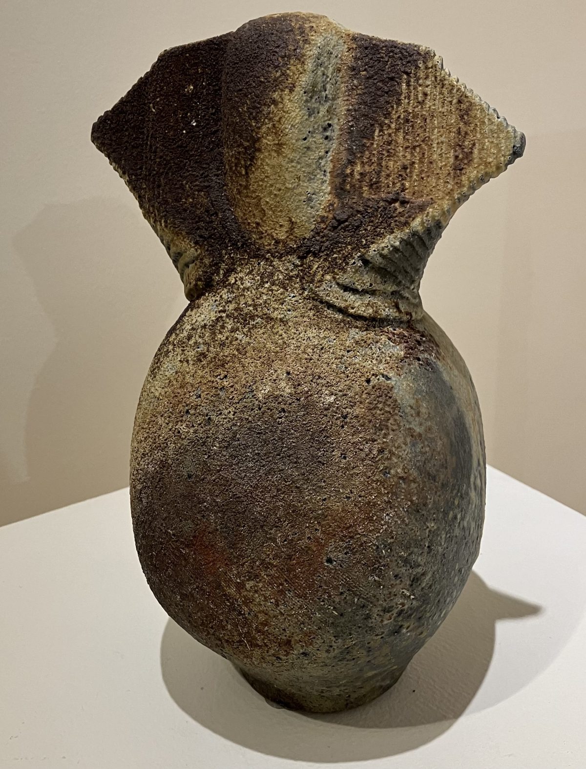 A textured ceramic vessel with earthy tones, featuring a bulbous base and a wide, angular top with subtle ridged details.