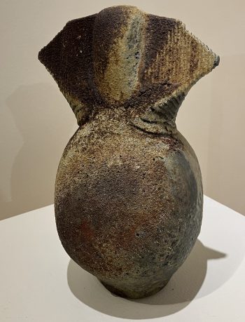 A textured ceramic vessel with earthy tones, featuring a bulbous base and a wide, angular top with subtle ridged details.