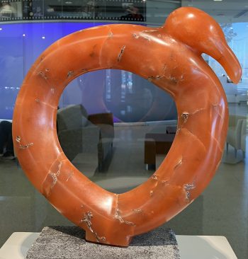 A sculpture of a circular orange alabaster bird with subtle veining, symbolizing wholeness and eternity, placed on a granite base in a modern interior setting.