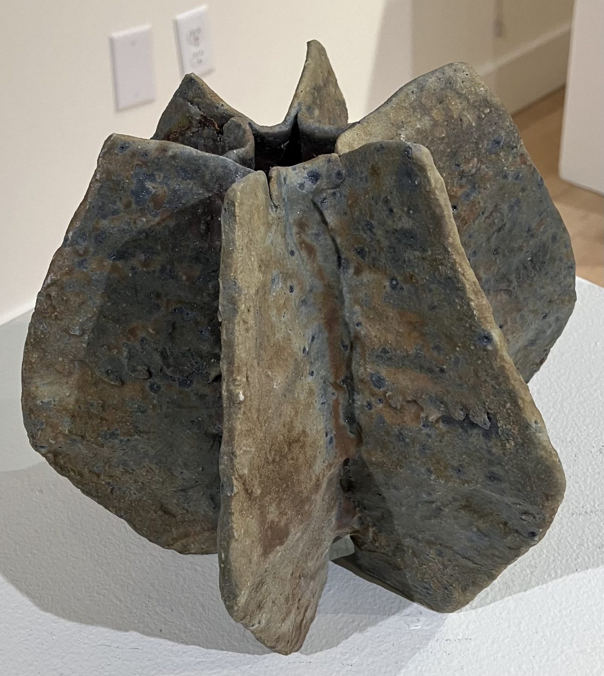An abstract ceramic sculpture with sharp, angular folds and earthy, textured surfaces, resembling an open flower or starburst.