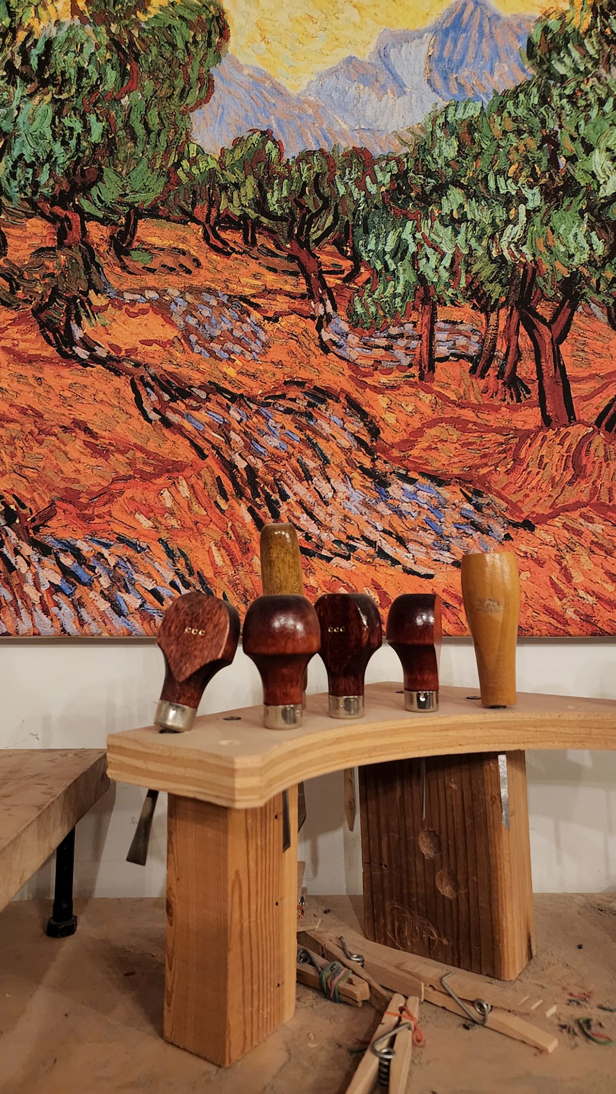 A collection of wooden-handled tools with polished finishes, arranged on a wooden holder, placed in front of a vibrant painting of a Van Gogh-style landscape with swirling colors and textured brushstrokes.