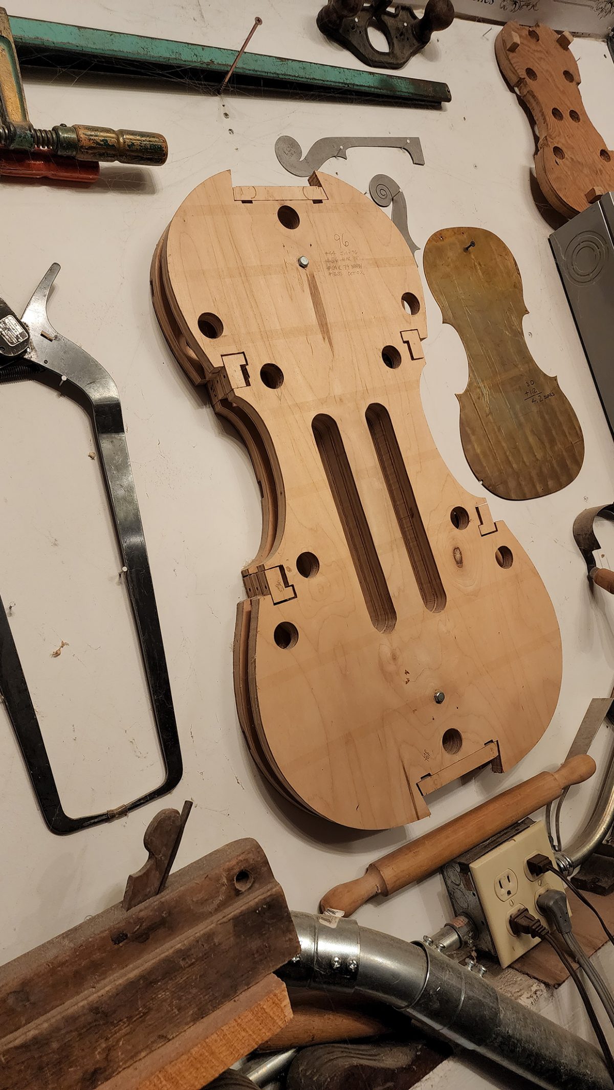 A wooden mold for violin-making, featuring precise cutouts and measurements, displayed on a workbench surrounded by tools like clamps, a saw, and other woodworking instruments.