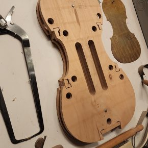 A wooden mold for violin-making, featuring precise cutouts and measurements, displayed on a workbench surrounded by tools like clamps, a saw, and other woodworking instruments.