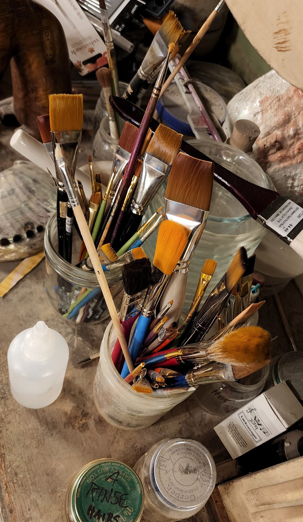 A collection of paintbrushes of various sizes and shapes stored in glass jars on a cluttered artist's workspace, surrounded by additional art supplies like jars, tubes, and bottles.
