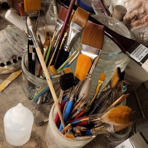 A collection of paintbrushes of various sizes and shapes stored in glass jars on a cluttered artist's workspace, surrounded by additional art supplies like jars, tubes, and bottles.