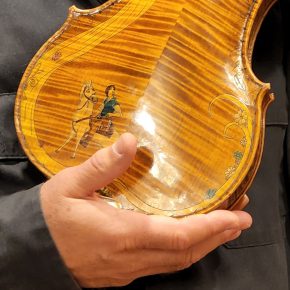 violin back plate with a richly figured maple pattern, decorated with painted illustrations of a person with a horse and floral embellishments, held by a person.