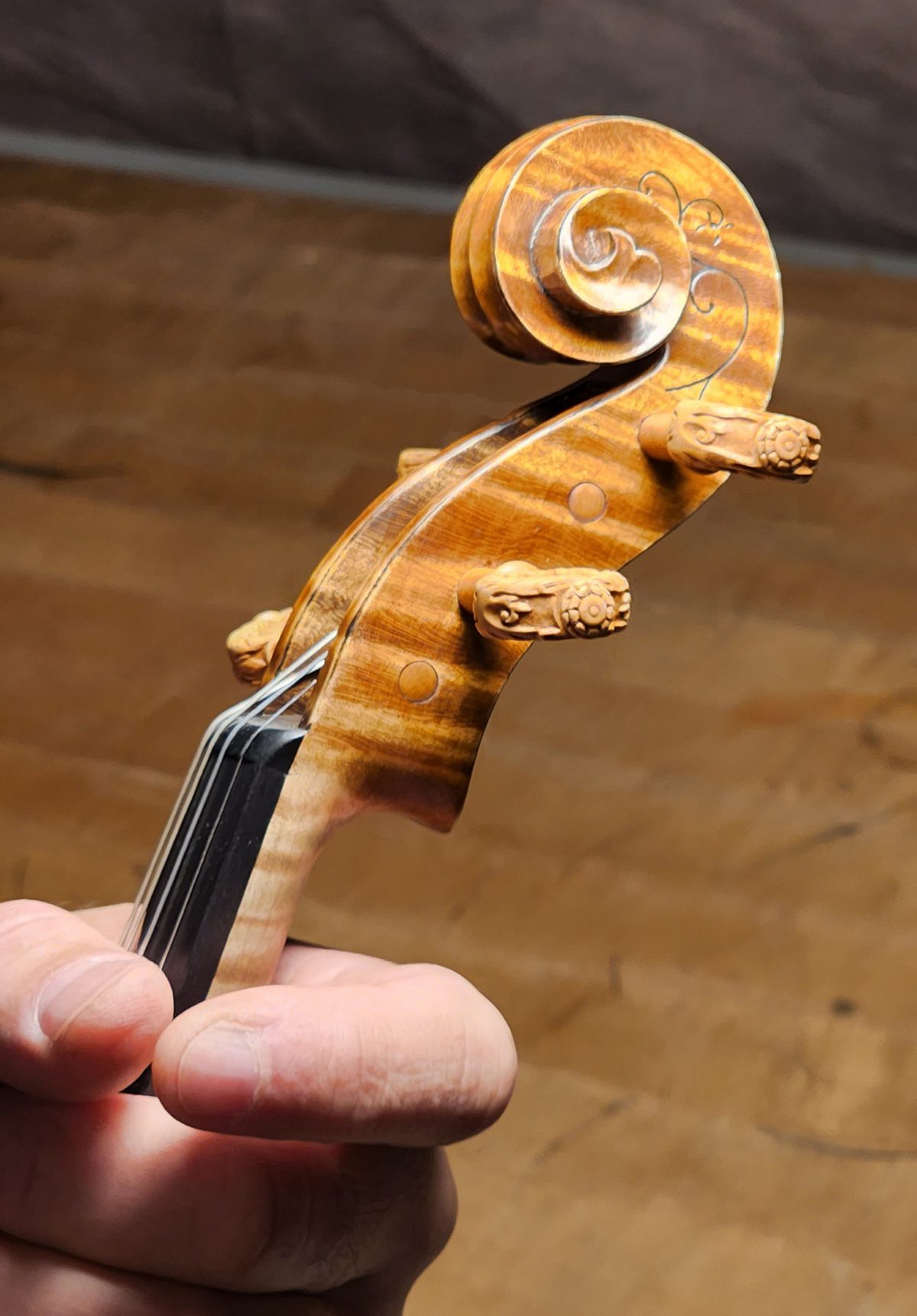 A detailed image of a violin scroll with ornately carved wooden pegs, held by a person’s hand against a wooden background.