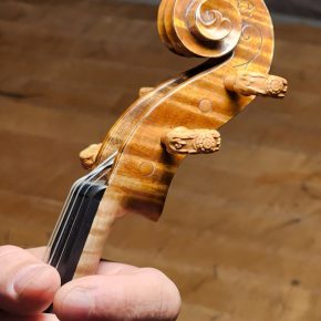 A detailed image of a violin scroll with ornately carved wooden pegs, held by a person’s hand against a wooden background.