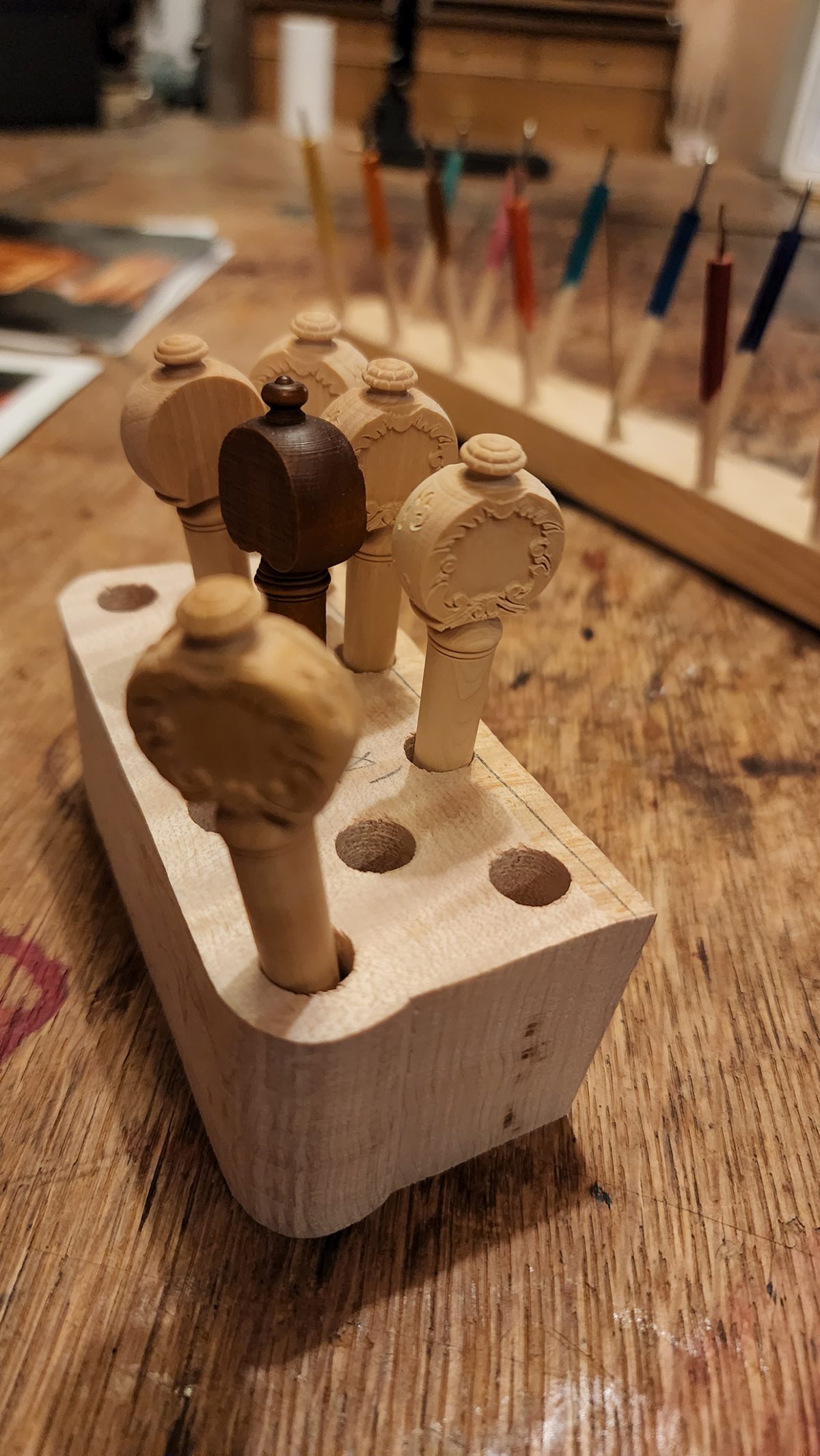 A close-up view of intricately carved wooden violin pegs displayed in a wooden holder, with colorful violin repair tools blurred in the background.