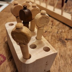 A close-up view of intricately carved wooden violin pegs displayed in a wooden holder, with colorful violin repair tools blurred in the background.