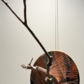 A mixed-media sculpture featuring a wooden branch extending outward from a circular base. The base is painted with abstract tree-like patterns in earthy tones and is adorned with orange string radiating outward in a fan-like arrangement, creating a dynamic interplay between natural and geometric elements.