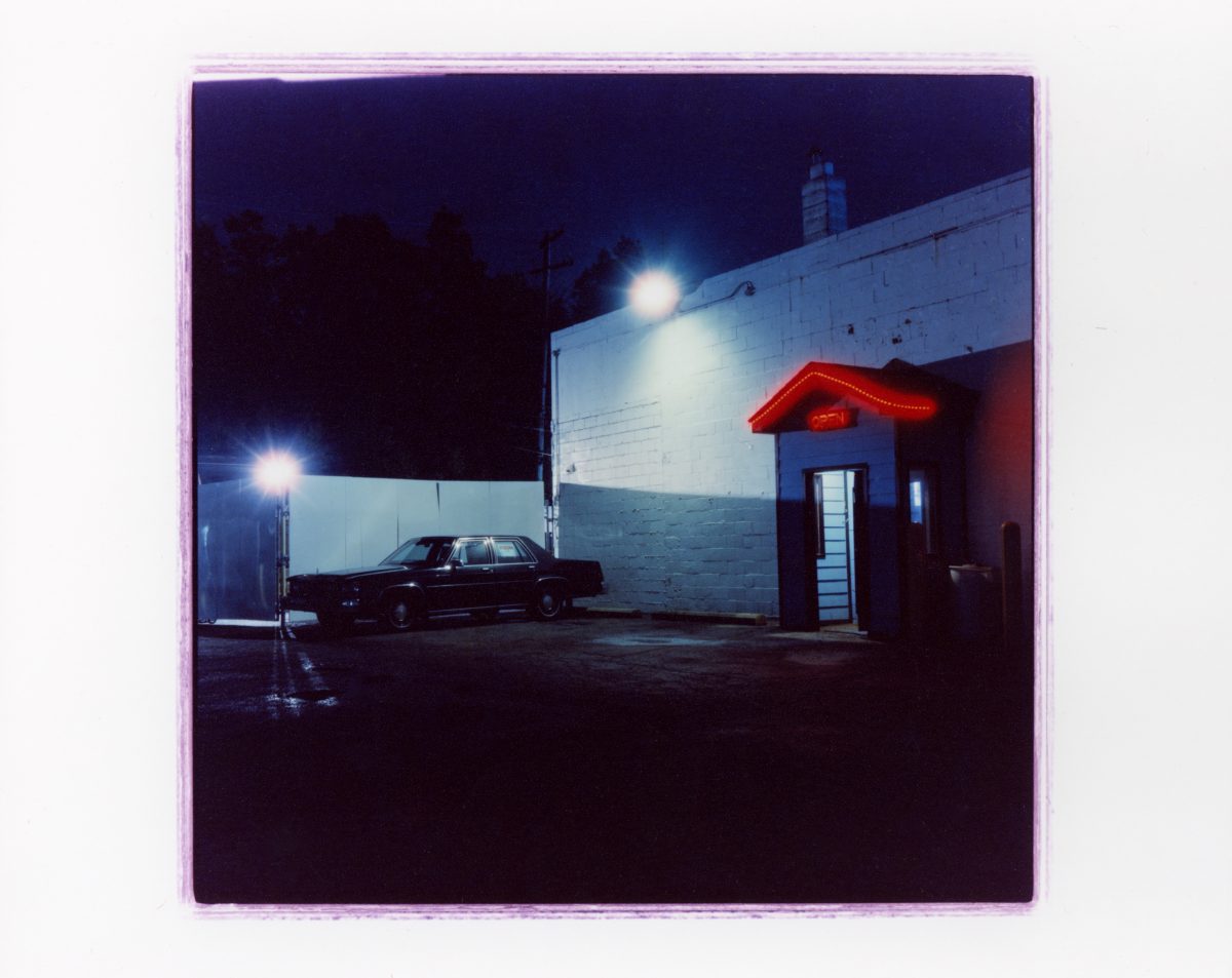 A nighttime photograph of a parking lot with a vintage car parked near a building, featuring a neon red sign reading 'OPEN' under a small awning.