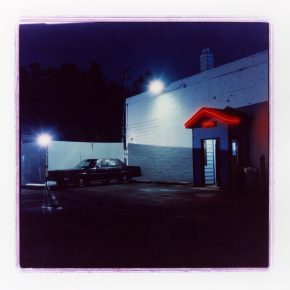 A nighttime photograph of a parking lot with a vintage car parked near a building, featuring a neon red sign reading 'OPEN' under a small awning.