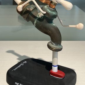 A whimsical sculpture titled "The Running Faucet" by Ron Christensen, depicting a faucet anthropomorphized as a running figure, complete with a blue cap, red shoes, and dynamic posture.