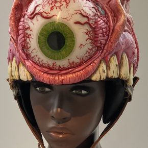 A striking sculptural helmet by William Robbins, designed as a grotesque, one-eyed monster with intricate detailing, including veiny textures, sharp teeth, and a vivid green eyeball.