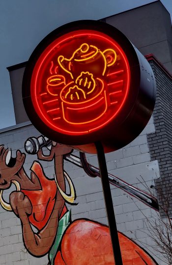 A round neon sign in red and yellow displaying a steaming tea kettle pouring into a cup, with a container of dumplings below, mounted against a wall with a mural of a singing woman.
