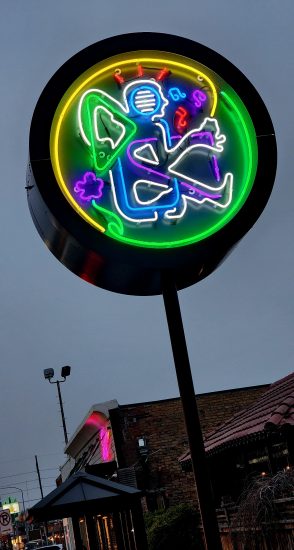 A round neon sign glowing in blue, green, and yellow, depicting a stylized humanoid figure with wings sitting cross-legged, surrounded by abstract shapes.