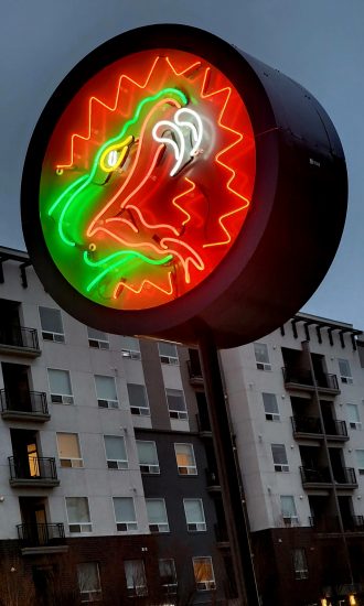 A circular neon sign depicting a red and green stylized snake head with white flames coming from its mouth, glowing against a modern apartment complex.