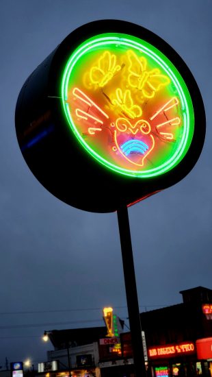 A round neon sign glowing in green, yellow, and red, featuring three butterflies above a heart with decorative swirls, against a dark sky and a row of city lights.