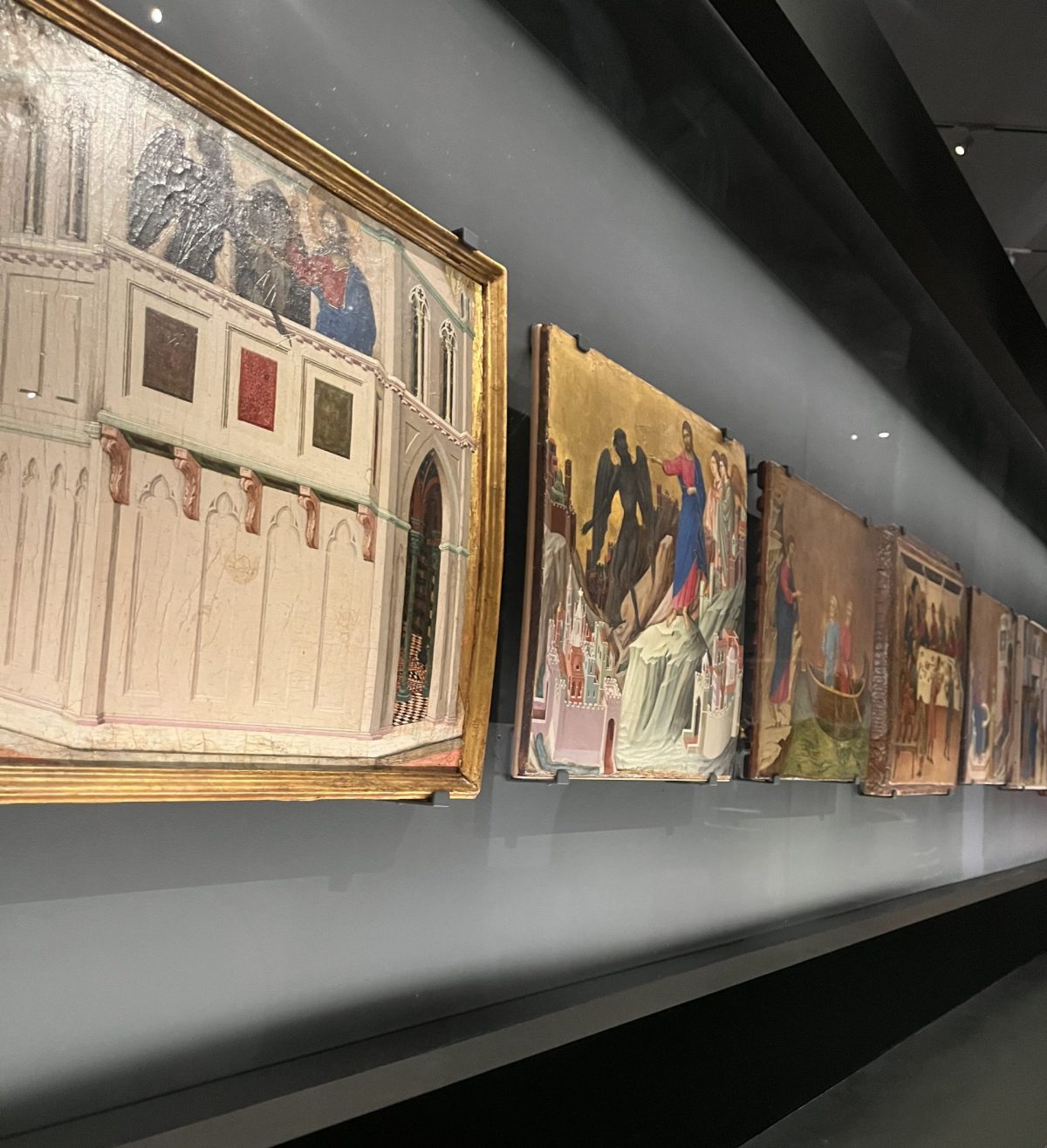 A display of medieval paintings in a museum, showing a row of illuminated religious-themed artworks with golden backgrounds.