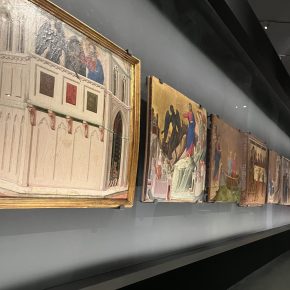 A display of medieval paintings in a museum, showing a row of illuminated religious-themed artworks with golden backgrounds.