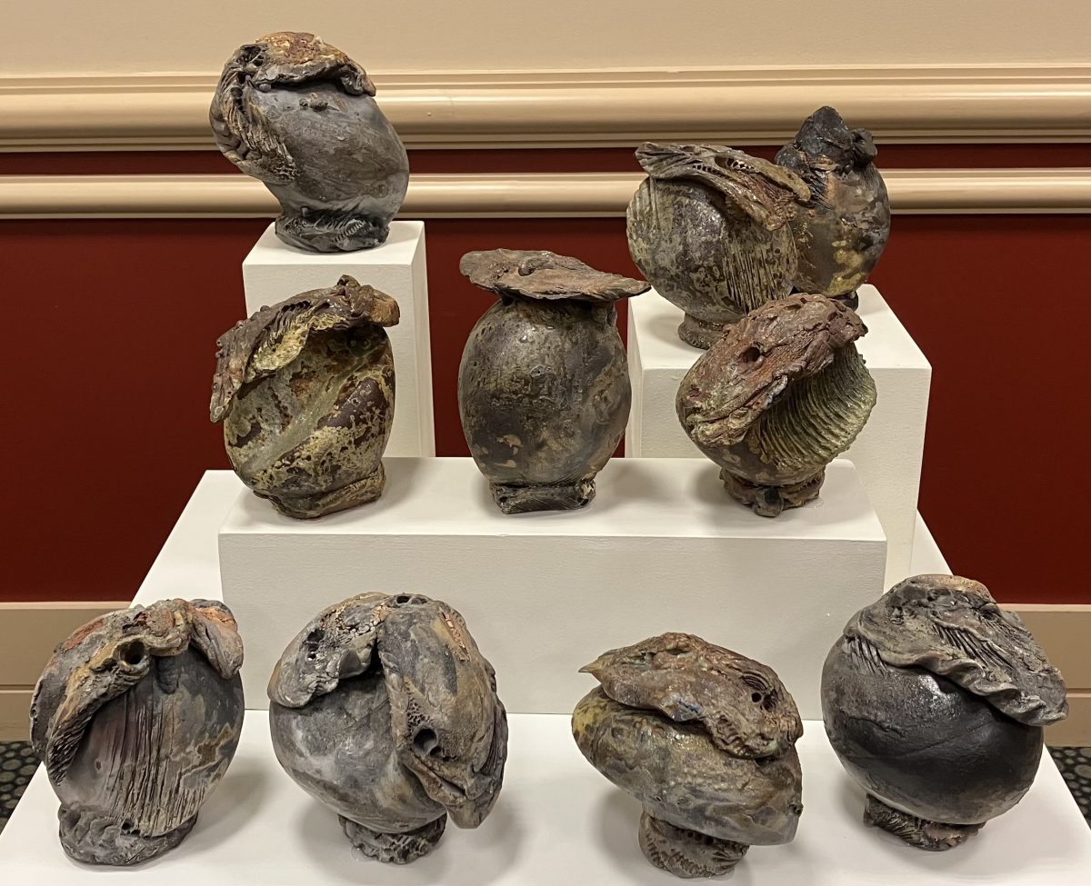 A group of organic-shaped ceramic vessels with rough, textured surfaces and earthy glazes, arranged on a tiered white pedestal.