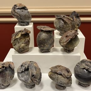 A group of organic-shaped ceramic vessels with rough, textured surfaces and earthy glazes, arranged on a tiered white pedestal.