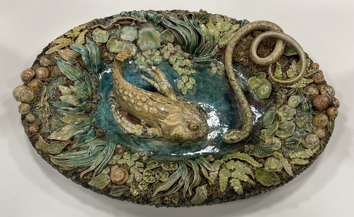An oval ceramic platter featuring a sculpted fish and snake surrounded by detailed foliage and snails, all rendered in earthy glazes.