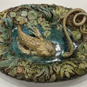 An oval ceramic platter featuring a sculpted fish and snake surrounded by detailed foliage and snails, all rendered in earthy glazes.