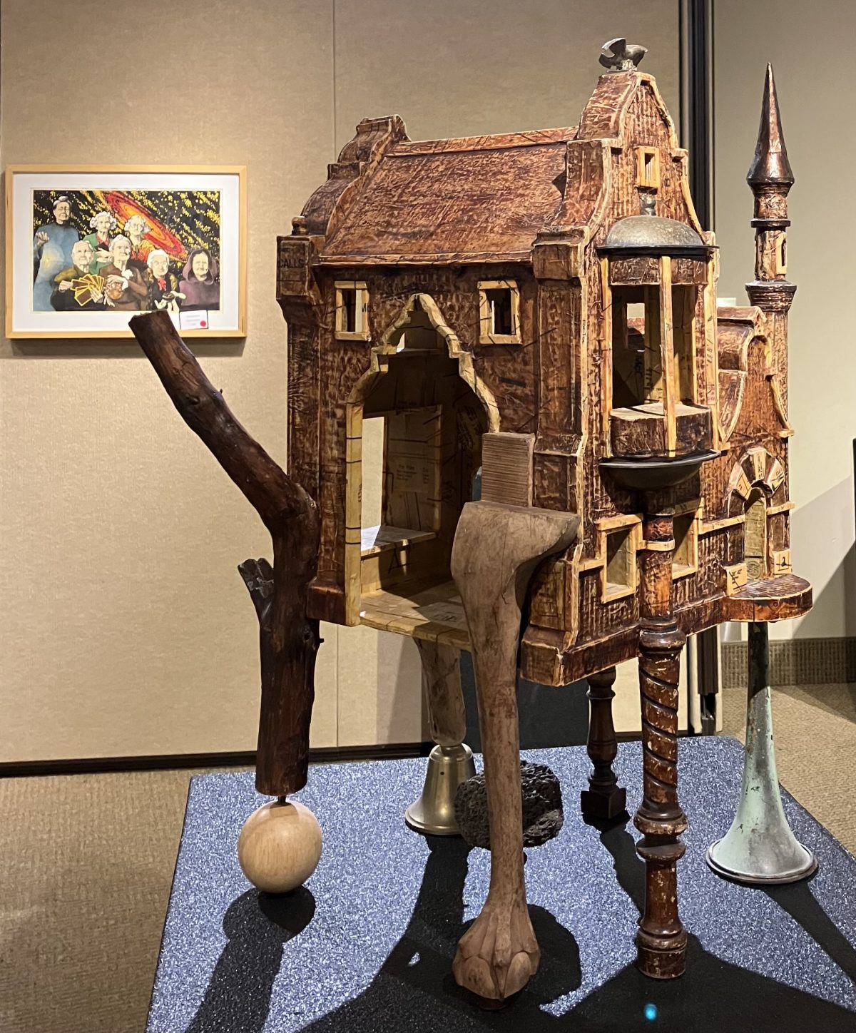 A detailed assemblage sculpture resembling a rustic house with a pointed turret, wooden textures, and mixed materials including metal elements. The structure appears to be supported by a carved animal leg, a wooden branch, and decorative objects resembling antique furniture.