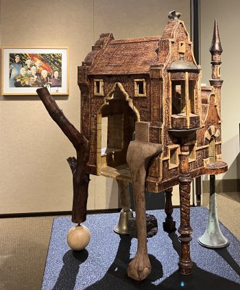 A detailed assemblage sculpture resembling a rustic house with a pointed turret, wooden textures, and mixed materials including metal elements. The structure appears to be supported by a carved animal leg, a wooden branch, and decorative objects resembling antique furniture.