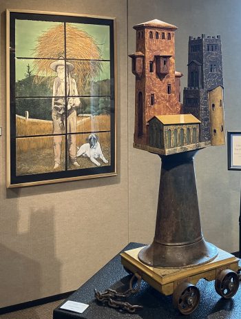 A sculptural piece featuring two miniature tower structures, one brown and one dark gray, positioned atop a large, cone-shaped metal base mounted on wooden wheels with chains attached.