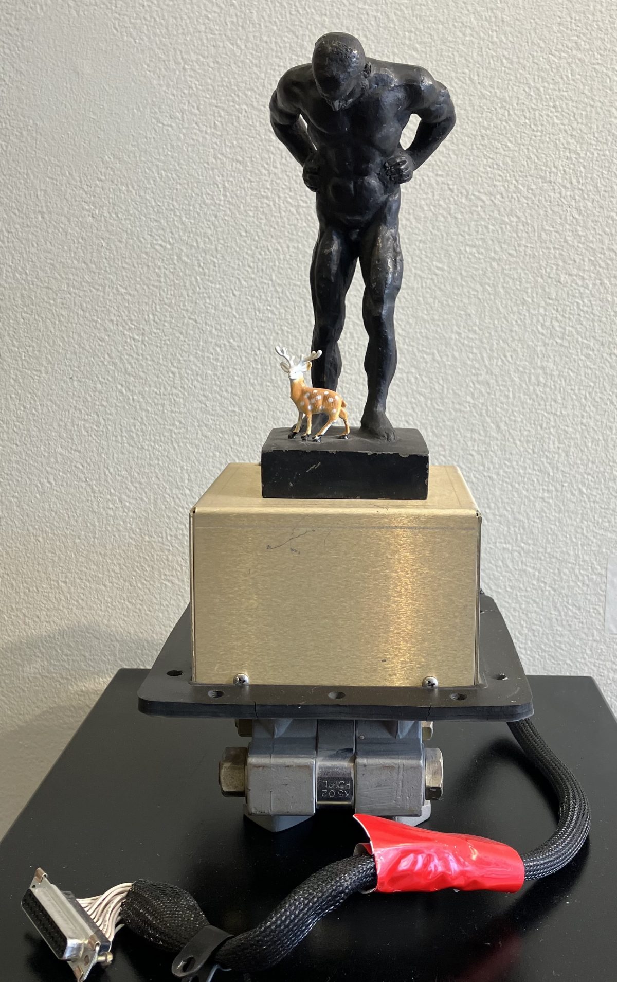 Assemblage sculpture of a black muscular figure leaning over a tiny deer, mounted on a gold metal box with electronic cables extending from the base.