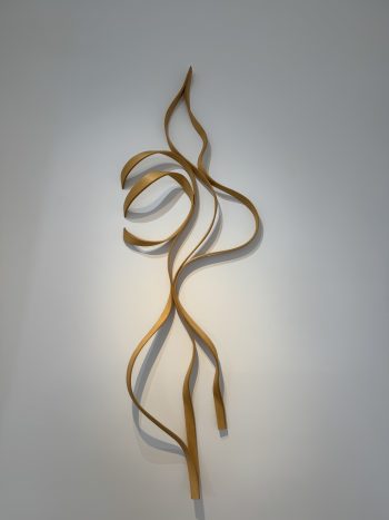 A delicate wooden wall sculpture in golden tones with flowing curves, resembling swirling flames or tendrils of smoke. The piece casts gentle shadows on the white wall behind it.