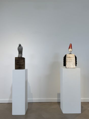 Two sculptures displayed on white pedestals: one features a minimalist, stone-like figure on a wooden base, while the other presents a stylized human bust with a red conical hat and a boxy wooden torso.