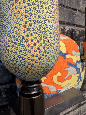 Close-up of a sculptural artwork featuring a yellow, dotted, pod-like form covered in multicolored circular patterns, mounted on a metallic base in front of a vividly patterned orange and blue backdrop.