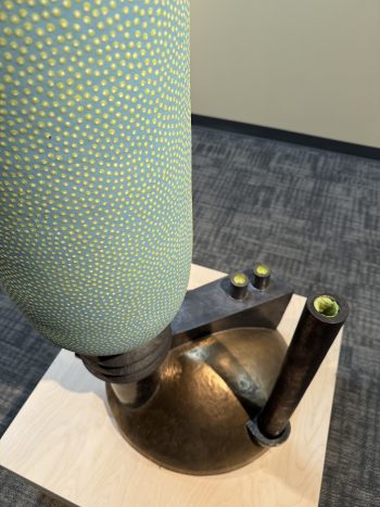 Green-and-yellow textured sculptures with black metallic bases, some wall-mounted) appears to be from a contemporary ceramic or mixed-media exhibition. The repetitive, pointillist surface treatment evokes a meditative or cellular feel. The contrast between the organic, patterned forms and industrial-looking mounts is especially striking.