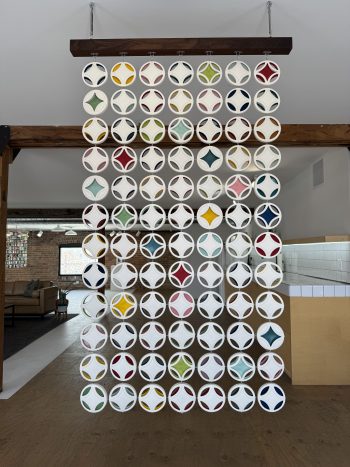 Wall-mounted installation featuring round, white ceramic elements with spiral cutouts, connected in a grid and suspended in front of a brick wall and window.