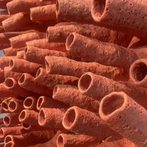 A close-up view of a ceramic sculpture composed of numerous porous, coral-like tubular structures in a vibrant, textured red-orange hue. The tubes extend outward, creating a sense of organic movement and density.