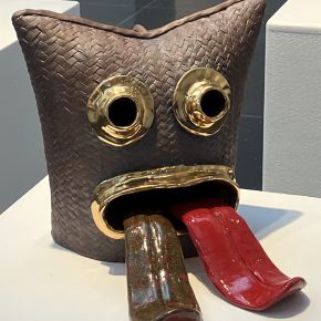 Sculptural ceramic mask with dark woven textures, exaggerated circular golden eyes, and a large mouth with two protruding, glossy red tongues.