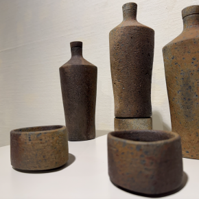 A collection of ceramic vessels in various earthy tones with textured surfaces, including bottles and small cups.