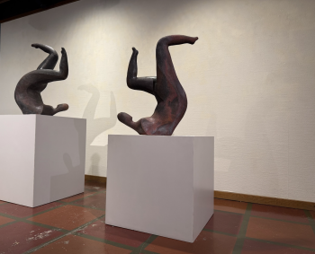 Two abstract ceramic sculptures of human figures in upside-down, contorted poses, displayed on white pedestals in a gallery setting.