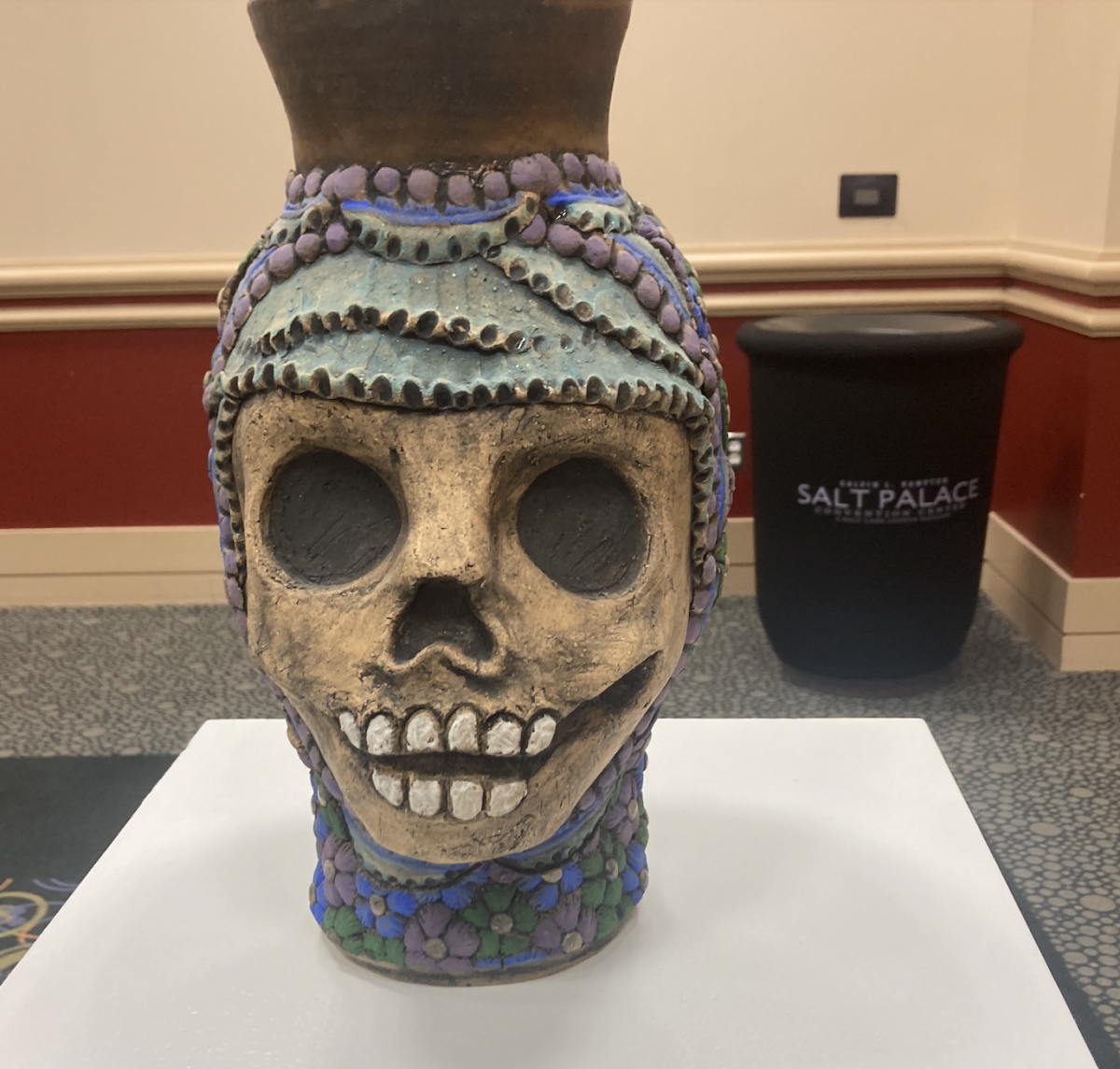 A ceramic vessel with a sculpted skull face and decorative beading in purple and turquoise hues, displayed on a pedestal in front of a “Salt Palace” branded container.