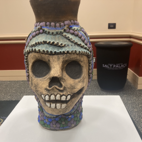 A ceramic vessel with a sculpted skull face and decorative beading in purple and turquoise hues, displayed on a pedestal in front of a “Salt Palace” branded container.