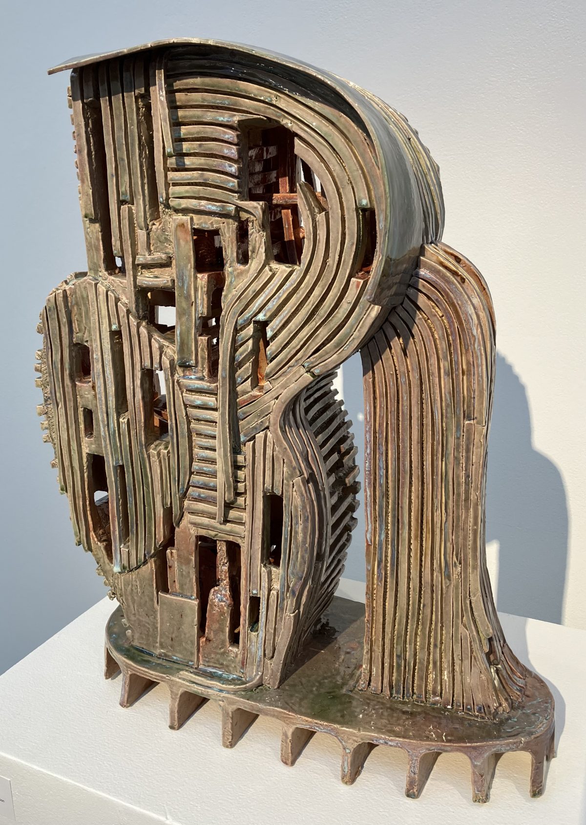 A large ceramic sculpture with an architectural quality, featuring stacked and curved layers forming an abstract structure. The piece has an earthy, metallic glaze with openings and intricate details.
