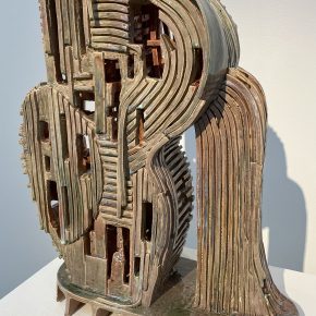 A large ceramic sculpture with an architectural quality, featuring stacked and curved layers forming an abstract structure. The piece has an earthy, metallic glaze with openings and intricate details.